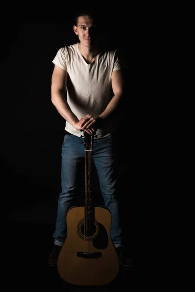 Guitarist, music. A young man stands with an acoustic guitar, on a black isolated background. Vertical frame — Stock Photo, Image