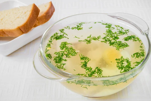 Tasty breakfast. Appetizing colorless soup with herbs in a transparent plate, bread in a white plate, on a white tablecloth. Horizontal frame — Stock Photo, Image