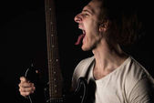 Hudba a kreativity. Pohledný mladý muž v tričku, s elektrickou kytarou, drží se jeho jazyk, na černém pozadí izolované. Vodorovný rámeček