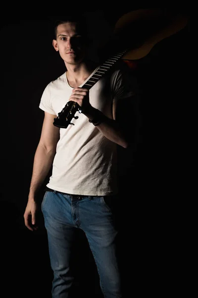 Music and creativity. Handsome young man in a T-shirt and jeans stands with an acoustic guitar on his shoulder, on a black isolated background. Vertical frame — Stock Photo, Image
