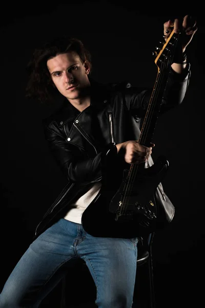 Music and creativity. Handsome young man in a T-shirt, jacket and jeans, with an electric guitar, on a black isolated background. Vertical frame — Stock Photo, Image
