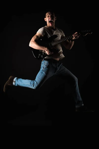 Music and creativity. Handsome young man in a T-shirt and jeans, jumping with an electric guitar, on a black isolated background. Vertical frame — Stock Photo, Image