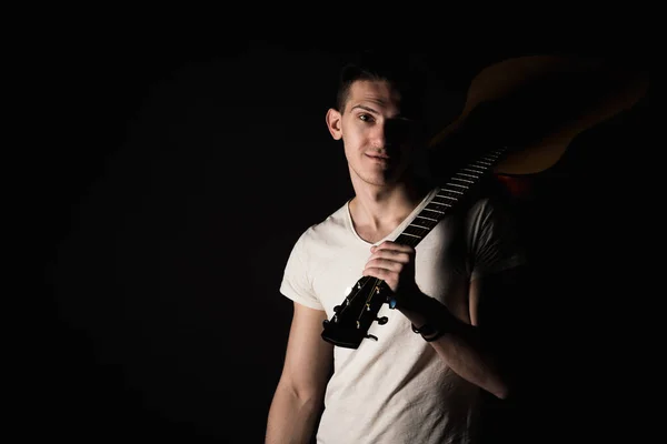 Music and creativity. Handsome young man in a T-shirt and jeans stands with an acoustic guitar on his shoulder, on a black isolated background. Horizontal frame — Stock Photo, Image