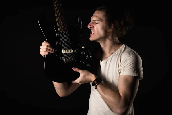 Music and creativity. Handsome young man in t-shirt, with electric guitar, showing teeth, on black isolated background. Horizontal frame — Stock Photo, Image