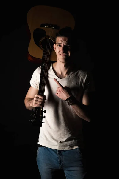 Música e criatividade. Um jovem bonito de t-shirt e jeans, com uma guitarra acústica no ombro, num fundo preto isolado. Quadro vertical — Fotografia de Stock