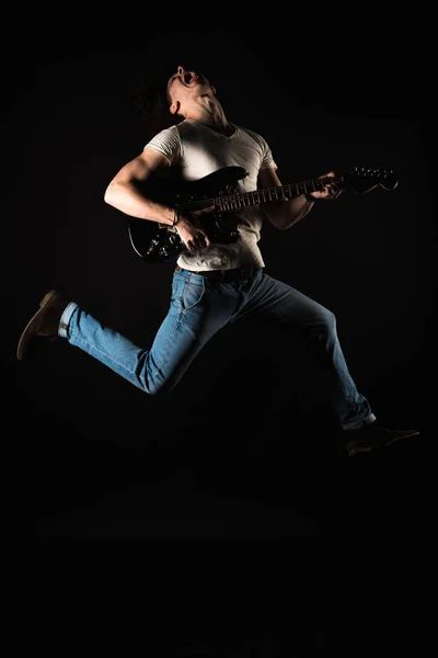 Musica e creatività. Bel giovanotto in maglietta e jeans, che salta con la chitarra elettrica, su uno sfondo nero isolato. Telaio verticale — Foto Stock