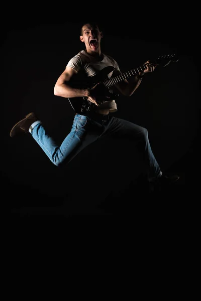 Music and creativity. Handsome young man in a T-shirt and jeans, jumping with an electric guitar, on a black isolated background. Vertical frame — Stock Photo, Image