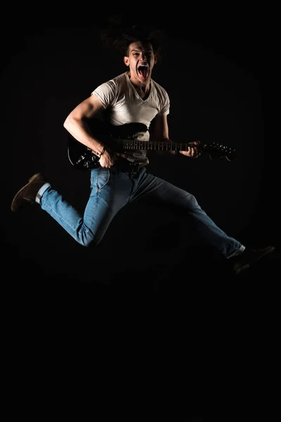 Music and creativity. Handsome young man in a T-shirt and jeans, jumping with an electric guitar, on a black isolated background. Vertical frame — Stock Photo, Image