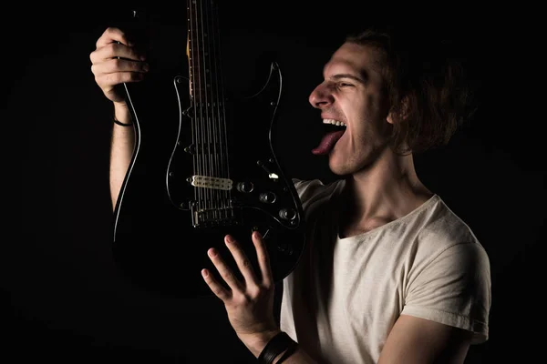 Hudba a kreativity. Pohledný mladý muž v tričku, s elektrickou kytarou, drží se jeho jazyk, na černém pozadí izolované. Vodorovný rámeček — Stock fotografie