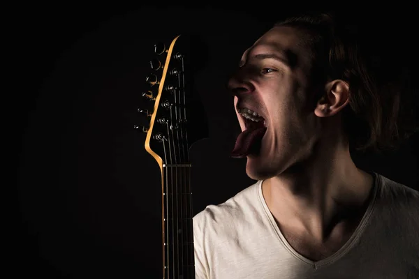 Music and creativity. Handsome young man in a T-shirt, with an electric guitar, sticks out his tongue, on a black isolated background. Horizontal frame — Stock Photo, Image