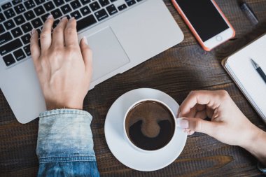 Üstten Görünüm tablosunun içen sabahları kahve masada bir fincan kahve, bir dizüstü bilgisayar ve cep telefonu ile ağ onu laptop sörf bir genç kızın üstünde. Yatay Çerçeve
