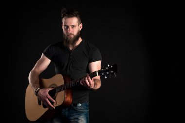 Sakallı karizmatik ve şık bir adam duruyor ve siyah bir izole arka planda akustik gitar çalar. Yatay Çerçeve