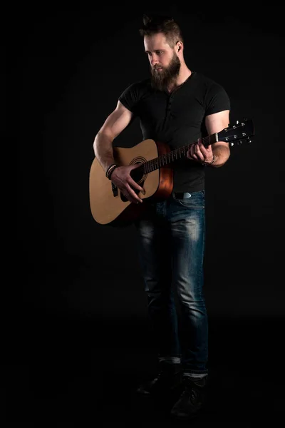 Um homem carismático e elegante com uma barba fica de comprimento total e toca uma guitarra acústica, em um fundo preto isolado. Quadro vertical — Fotografia de Stock