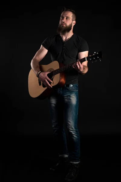 Ένας χαρισματικός και κομψό άνδρας με μούσι στέκεται ολόσωμο και παίζει μια ακουστική κιθάρα, πάνω σε μαύρο φόντο απομονωμένες. Κατακόρυφο πλαίσιο — Φωτογραφία Αρχείου