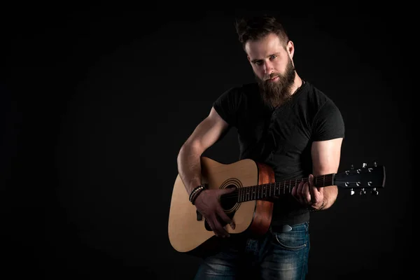 Un hombre carismático y elegante con barba se para y toca una guitarra acústica sobre un fondo negro aislado. Marco horizontal — Foto de Stock