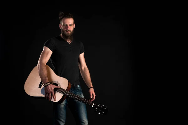 Um homem carismático e elegante com uma barba fica com uma guitarra acústica em um fundo preto isolado. Quadro horizontal — Fotografia de Stock