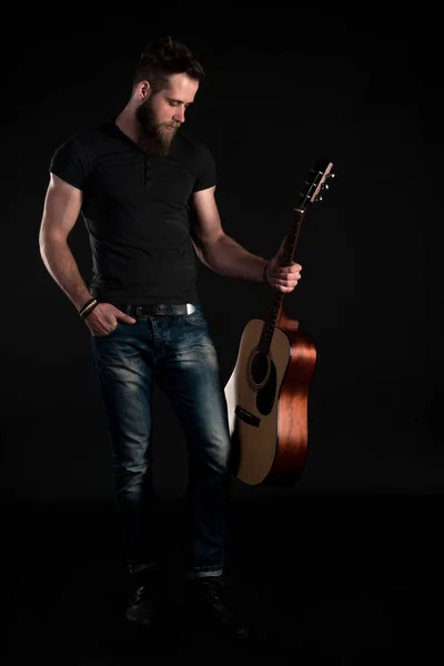Un uomo carismatico ed elegante con la barba si erge a figura intera con una chitarra acustica su uno sfondo nero isolato. Telaio verticale — Foto Stock