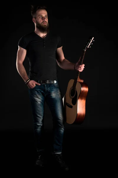 Um homem carismático e elegante com uma barba fica de comprimento total com uma guitarra acústica em um fundo preto isolado. Quadro vertical — Fotografia de Stock