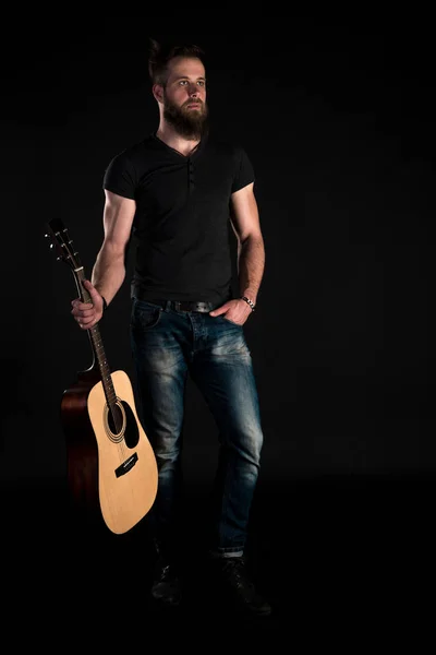 Un uomo carismatico ed elegante con la barba si erge a figura intera con una chitarra acustica su uno sfondo nero isolato. Telaio verticale — Foto Stock