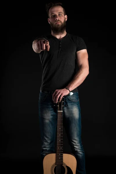 Un uomo carismatico ed elegante con la barba si erge a figura intera con una chitarra acustica su uno sfondo nero isolato. Telaio verticale — Foto Stock