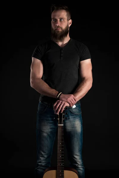 Харизматичний і стильний чоловік з бородою стоїть на повній довжині з акустичною гітарою на чорному ізольованому тлі. Вертикальна рамка — стокове фото