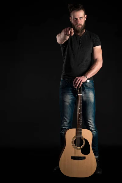 Um homem carismático e elegante com uma barba fica de comprimento total com uma guitarra acústica em um fundo preto isolado. Quadro vertical — Fotografia de Stock