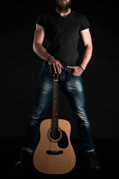 Um homem carismático e elegante com uma barba fica de comprimento total com uma guitarra acústica em um fundo preto isolado. Quadro vertical — Fotografia de Stock