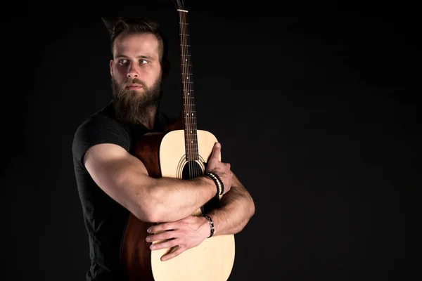 Un uomo carismatico ed elegante con la barba si erge a figura intera con una chitarra acustica su uno sfondo nero isolato . — Foto Stock