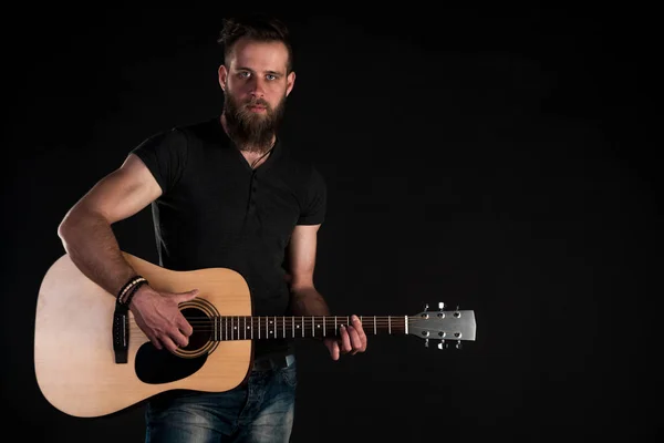 Un uomo carismatico ed elegante con la barba si erge a figura intera con una chitarra acustica su uno sfondo nero isolato . — Foto Stock