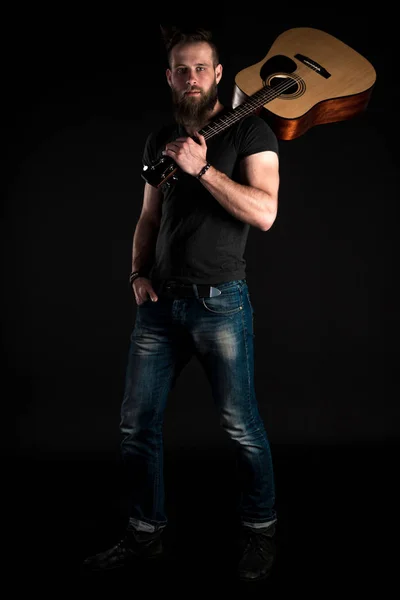 Um homem carismático e elegante com uma barba fica de comprimento total com uma guitarra acústica em um fundo preto isolado . — Fotografia de Stock