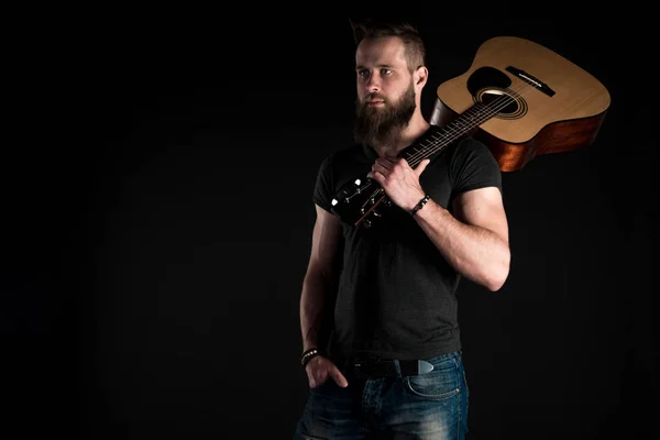 Un uomo carismatico ed elegante con la barba si erge a figura intera con una chitarra acustica su uno sfondo nero isolato . — Foto Stock