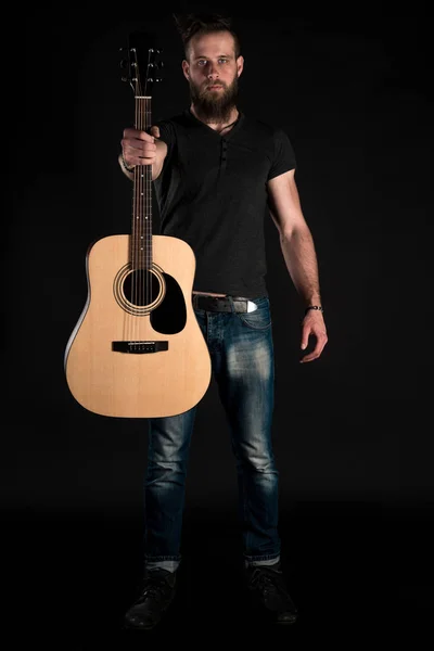 Um homem carismático e elegante com uma barba fica de comprimento total com uma guitarra acústica em um fundo preto isolado . — Fotografia de Stock