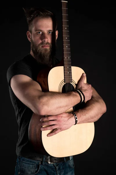 Um homem carismático e elegante com uma barba fica de comprimento total com uma guitarra acústica em um fundo preto isolado . — Fotografia de Stock