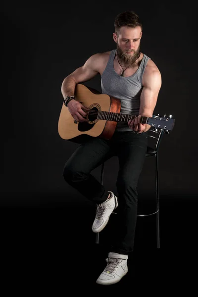 Um homem carismático com uma barba, tocando uma guitarra acústica, em um fundo preto isolado. Quadro vertical — Fotografia de Stock