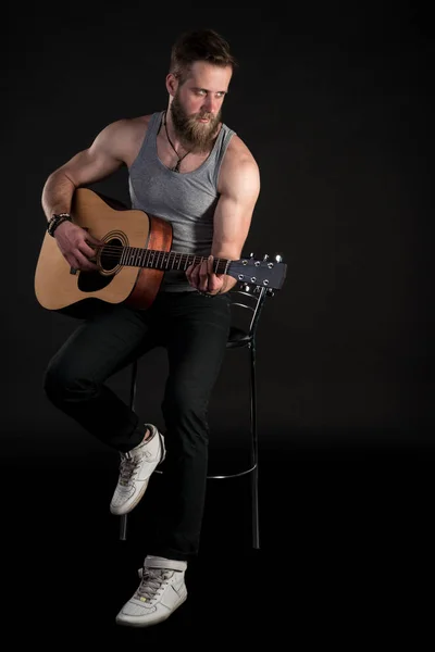 Um homem carismático com uma barba, tocando uma guitarra acústica, em um fundo preto isolado. Quadro vertical — Fotografia de Stock