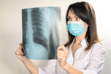 Koronavirüs ve güvenlik konusu. Hafif bir arka planda ameliyat kıyafeti giymiş genç bir kadın doktor röntgeni inceliyor..