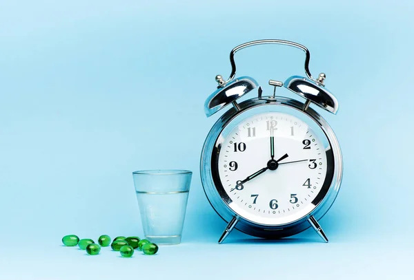 Medical green capsule pills, glass water and alarm clock on blue background. Concept health