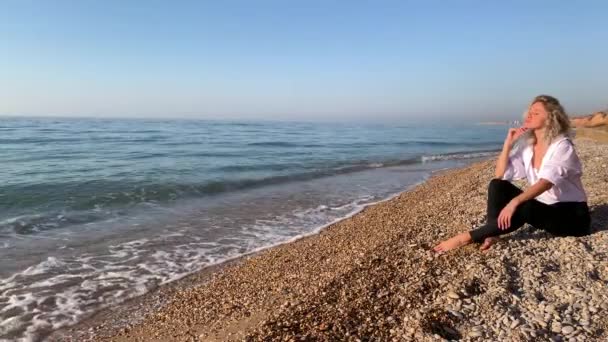 美しいブロンドの女性が座って 青い海の波の近くのビーチで瞑想 クリミア セヴァストポリ — ストック動画