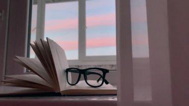 Pencerenin kenarında açık bir kitap duruyor, pencerenin dışında günbatımı var, yanında da okuma gözlüğü var. Yavaş çekim.