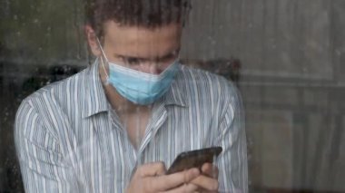 Yüz maskesi takan, COVID-19 hastası, hastanede karantinaya alınmış, telefonunu kullanan genç ve korkmuş bir adam.