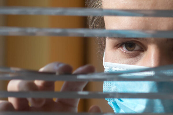 Молодой человек в медицинской маске смотрит в окно сквозь жалюзи. Карантин во время эпидемии
.