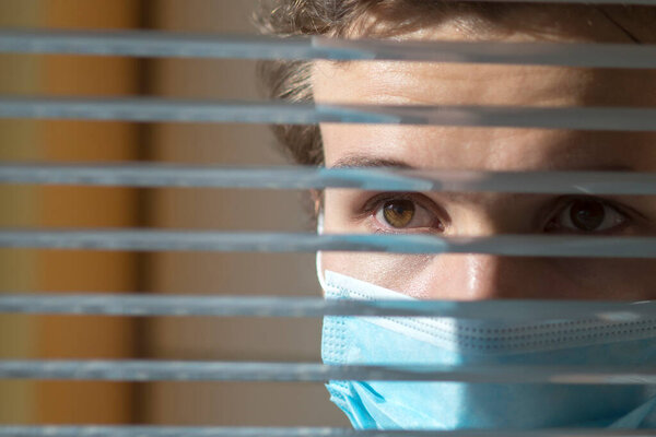 Молодой человек в медицинской маске смотрит в окно сквозь жалюзи. Карантин во время эпидемии
.