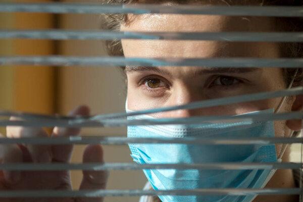 Молодой человек в медицинской маске смотрит в окно сквозь жалюзи. Карантин во время эпидемии
.