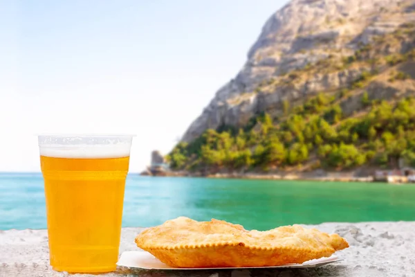 夏日清澈的背景下 在大海和山地海岸的背景下 杯子里放着啤酒和奶酪 放在纸盘上 免版税图库图片