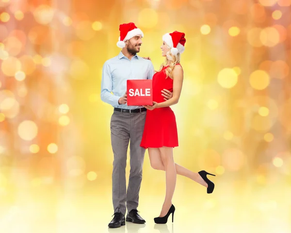 Casal feliz em chapéus de santa com sinal de venda vermelho — Fotografia de Stock