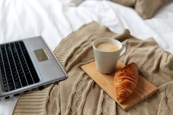 Ноутбук, кофе и круассан на кровати в уютном доме — стоковое фото