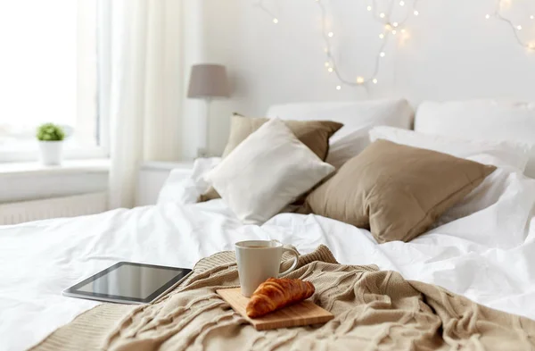 Tablet pc, coffee cup and croissant on bed at home — Stock Photo, Image