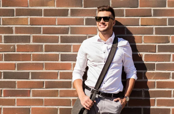 Молодой человек в солнцезащитных очках с сумкой на кирпичной стене — стоковое фото