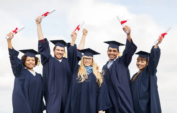 Happy students in mortar boards waving diplomas — Stock Photo, Image