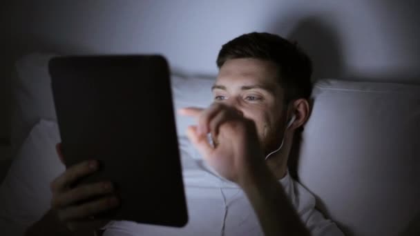 Muž s tablet pc a sluchátka v posteli v noci — Stock video